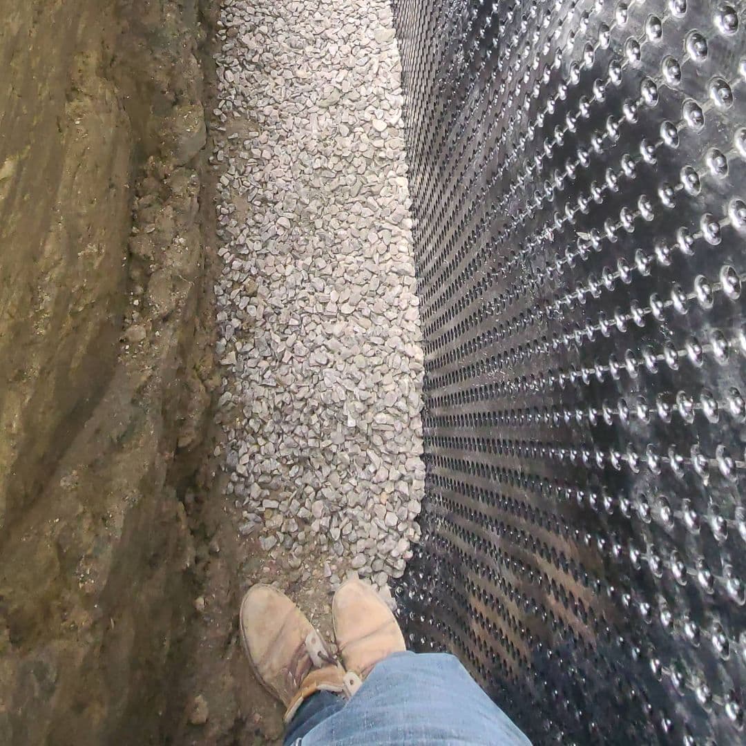 Close-up view of a foundation wall with dimpled waterproofing membrane installed, surrounded by gravel drainage layer and soil trench, with worker boots visible.