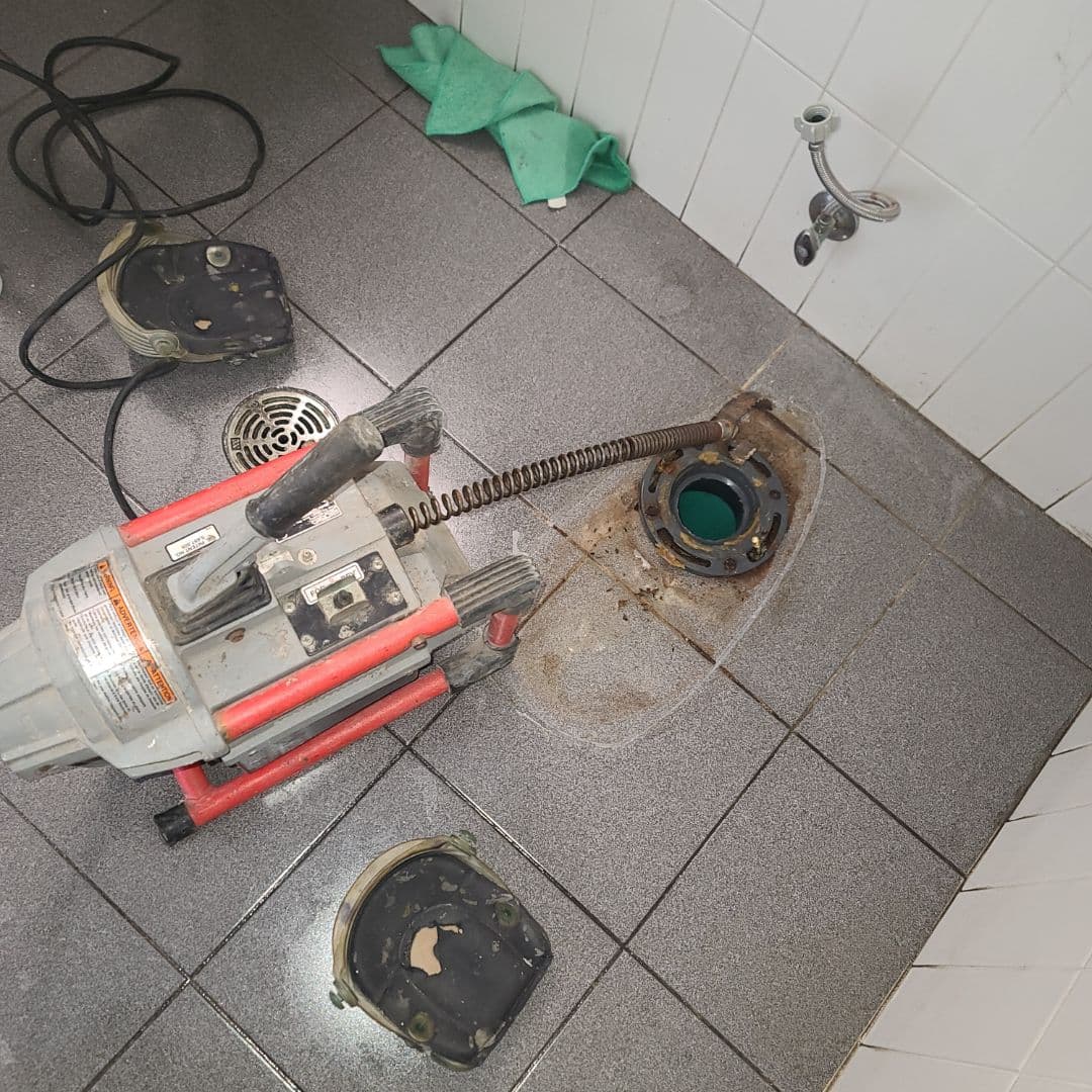 Drain machine with its cable in an open toilet flange on a tiled bathroom floor.
